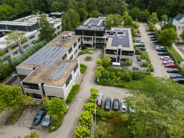 Luchtfoto van het gebouw aan de Burgemeester Verderlaan met zonnepanelen op het dak en omliggende parkeerplaatsen.